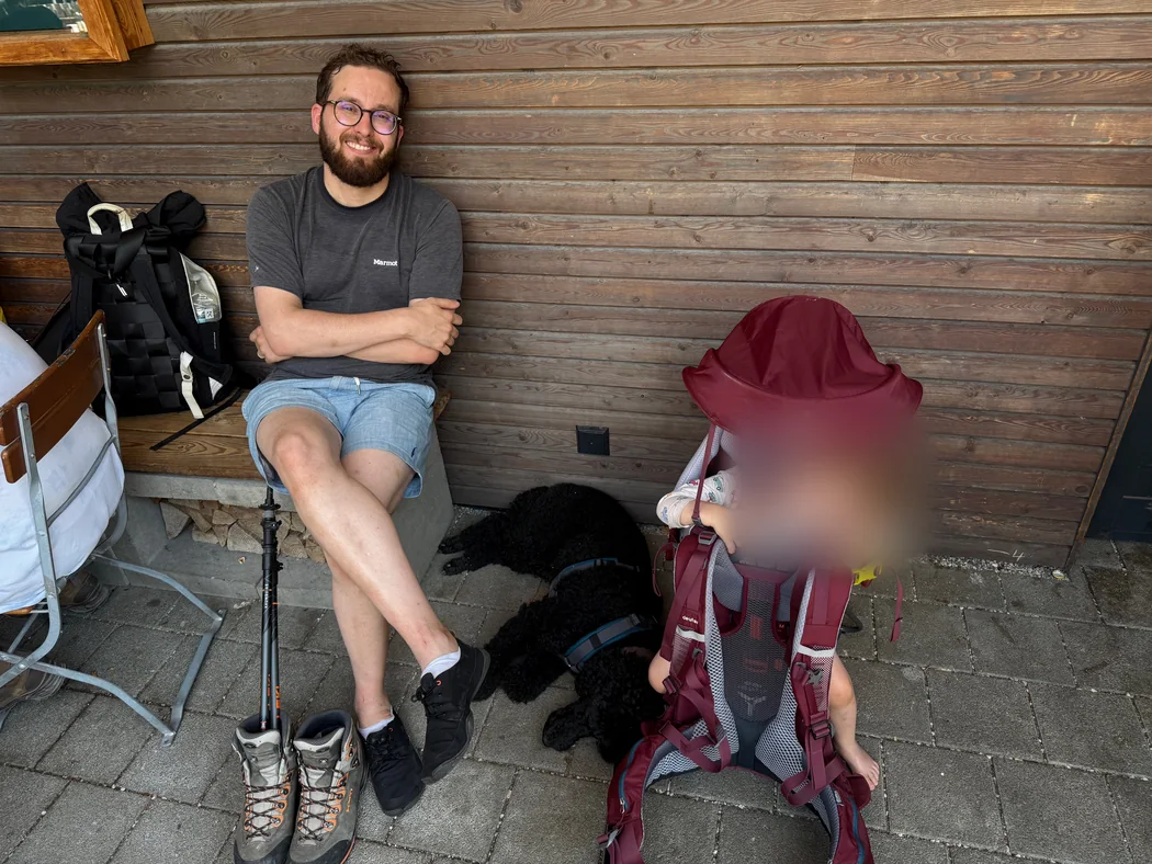 Seamus sitzt vor einer Hütte, Tiffany liegt neben ihm im Schatten
